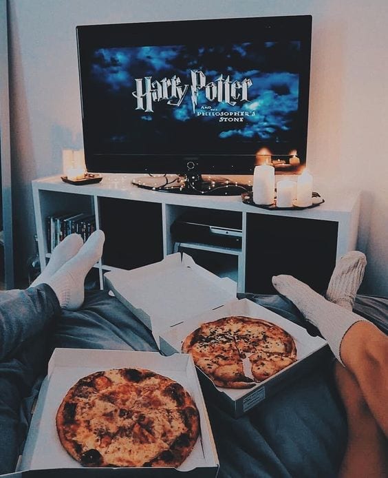 Casal sentado na cama, apenas as pernas aparecendo, com duas pizzas sobre a cama e a tv ligada com o filme do Harry Potter.