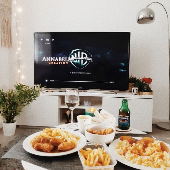 Mesa de centro, com batata frita, frango frito e taça de vinho e tv ao fundo com o logo da WB.