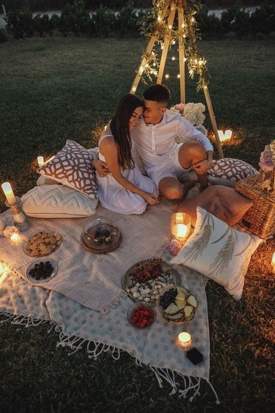 Casal sentado sob uma toalha branca em cima da grama, com almofadas em volta, com luzes de led e vela iluminando, com comidas para fondue.