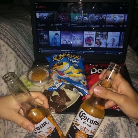 Casal deitados, brincando duas cervejas da marca Corona, com salgadinhos sobre o Notebook ligado com Netflix.