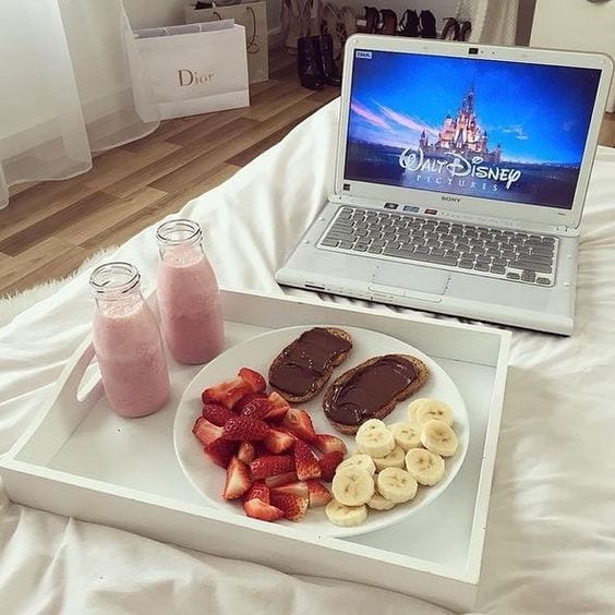 Bandeja branca sobre a cama, com um prato com panquecas, banana e morango cortado, dois copos com vitaminas de morango ao lado e o notebook a frente com o logo da disney.