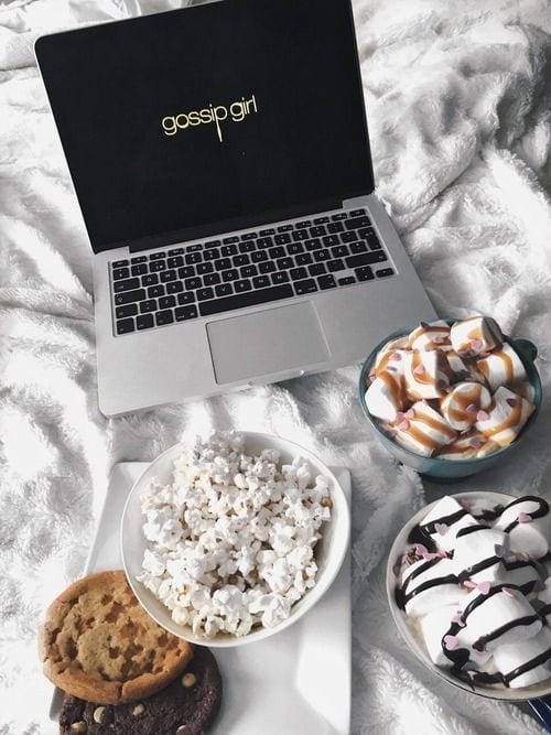 Notebook sobre a cama com o logo da série Gossip Girls com vasilha de pipoca, coockies e sorvete.
