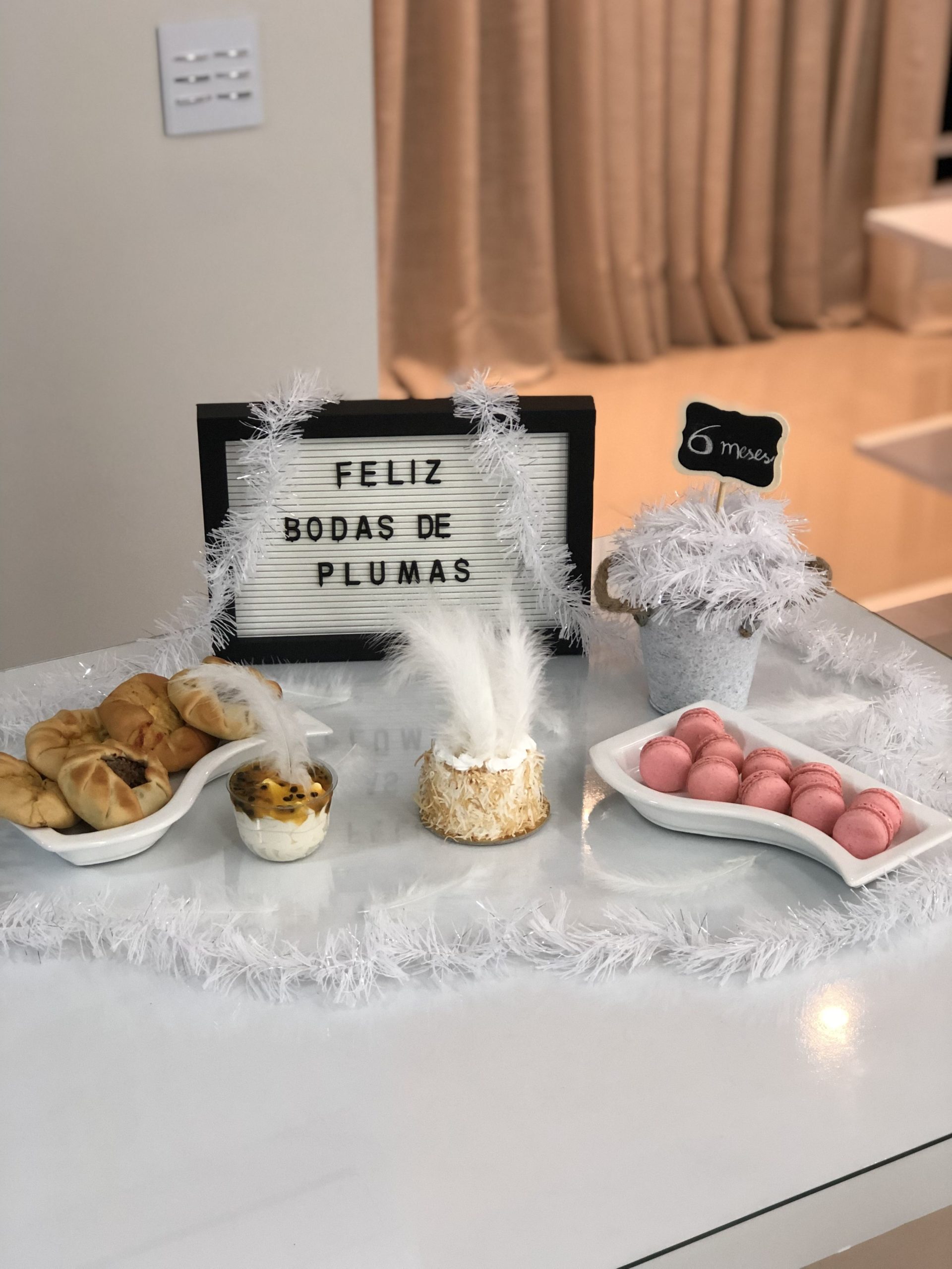 Mesa branca de vidro, com plumas em cima, placa decorativa branca escrito feliz bodas de plumas, e algumas esfihas, macorrones rosas e mousse de maracuja.