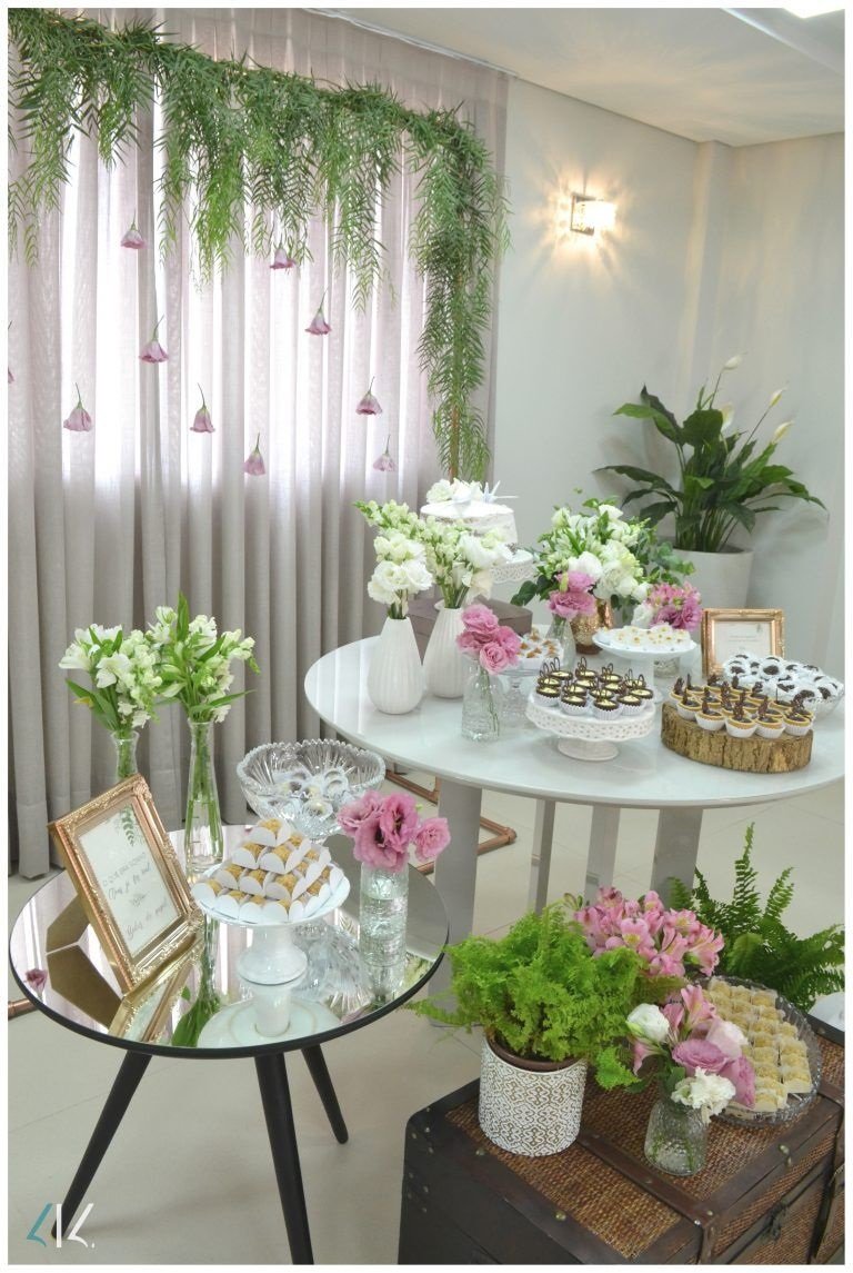 Duas mesas pequenas, decoradas com docinhos e vasos de vidro transparente com flores brancas e rosas. Cortina branca no fundo, com folhas verdes e flores rosas. Baú a frente das mesas com mais plantinhas e flores brancas e rosas.
