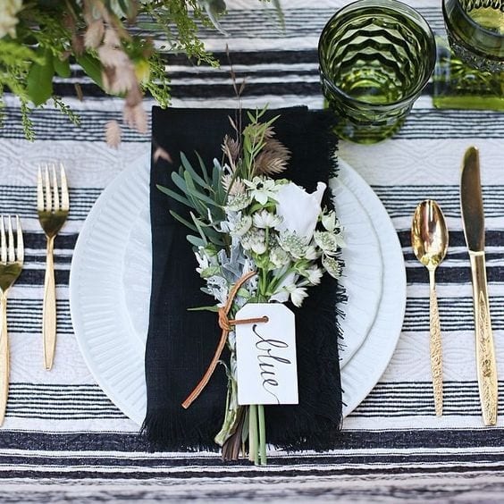 Mesa posta com pano branco com listras pretas, talheres dourados, pratos brancos, com um guardanapo preto e flores do campo por cima.