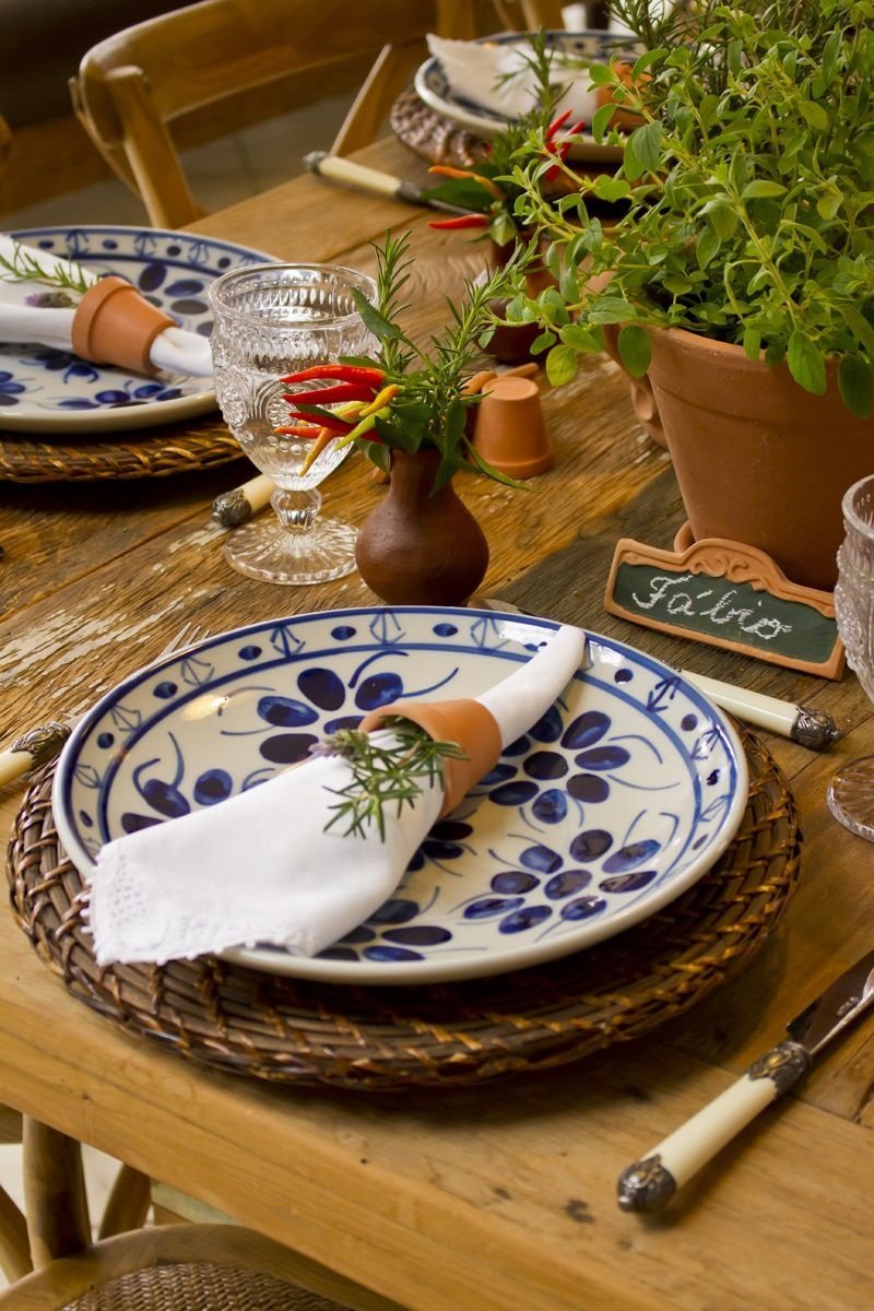 Mesa posta com pratos brancos e azuis, talheres em prata com o cabo branco e prata, guardanapo branco com alecrim acima do prato e vazo com algumas pimentas vermelhas e amarelas ao lado.