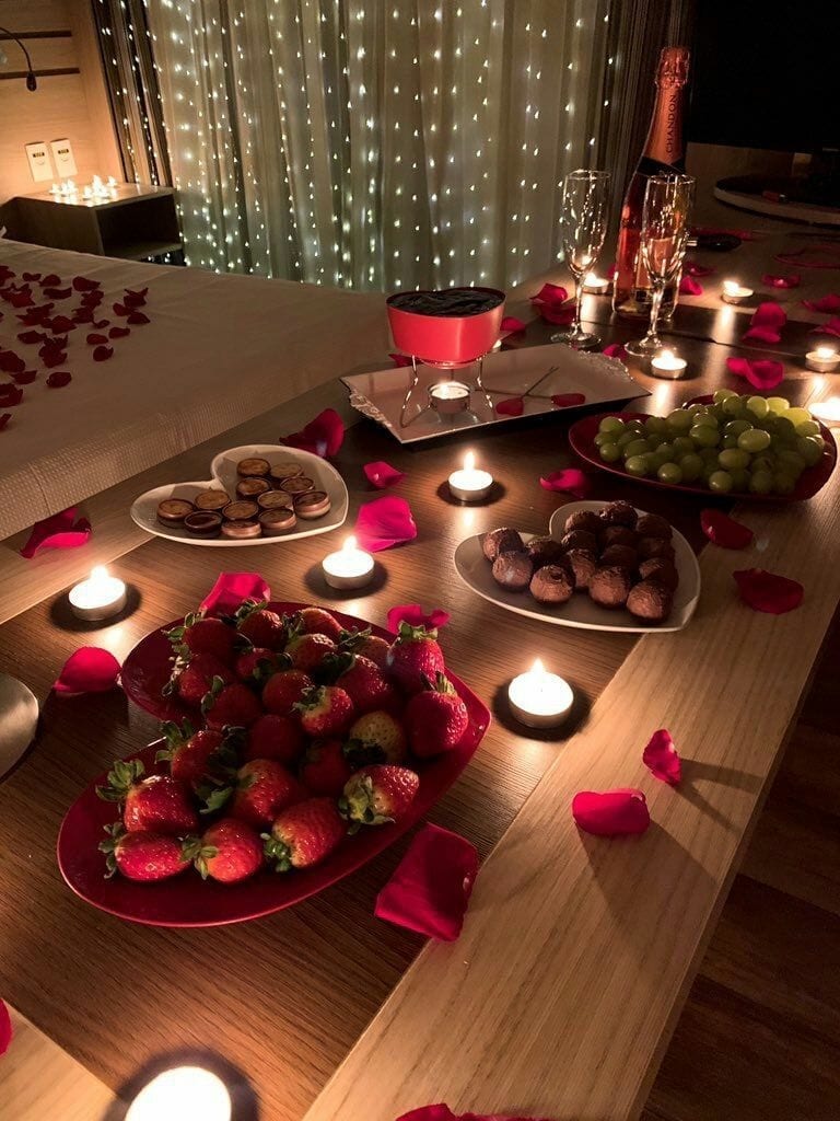 Mesa retangular de madeira marrom clara, pratos em forma de coração vermelho e branco, com frutas e chocolate, velas pequenas e pétalas de rosa sob a mesa.