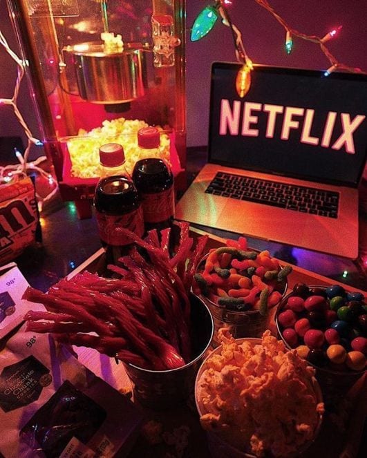 Fini, Pipoca e outros snacks sobre a mesa, com coca cola e m&m a esquerda, e notebook com a tela da Netflix.