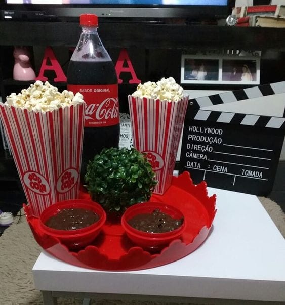 Mesa branca, com 2 sacos de pipoca branco e vermelho, uma garrafa de coca cola de 600ml e duas cumbucas pequenas de brigadeiro de colher.