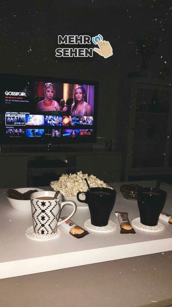 Mesa branca, com 3 canecas, pipoca e cockies e tv ao fundo ligada na Netflix.
