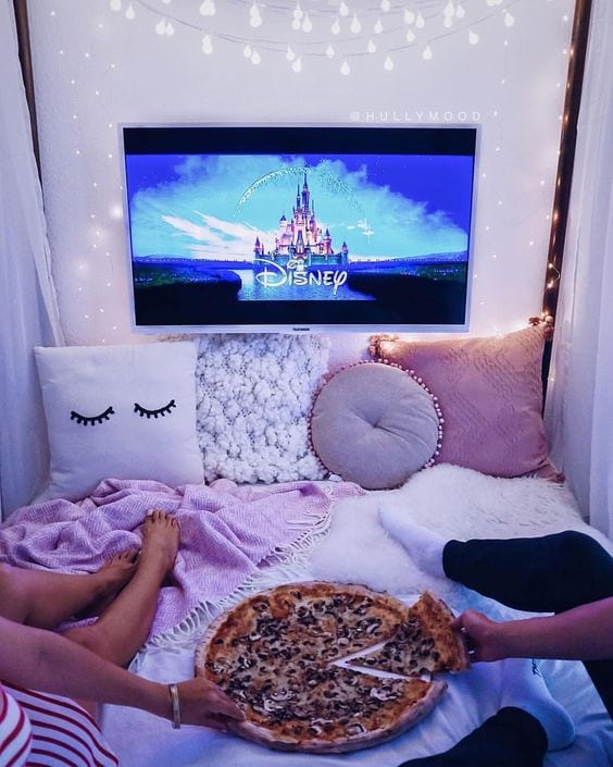 Amigas sentadas na cama, apenas as pernas aparecendo, com uma pizza entre elas na cama e a tv ligada com a imagem do castelo da disney.