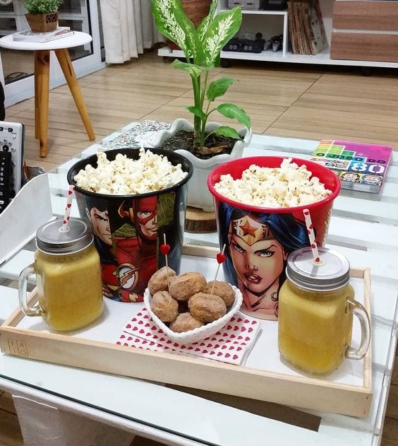 Bandeja branca, sobre a mesa de centro da sala, com um balde de pipoca pequeno vermelho da mulher maravilha e outro da liga da justiça preto, com pipoca, e copos amarelos com canudo.