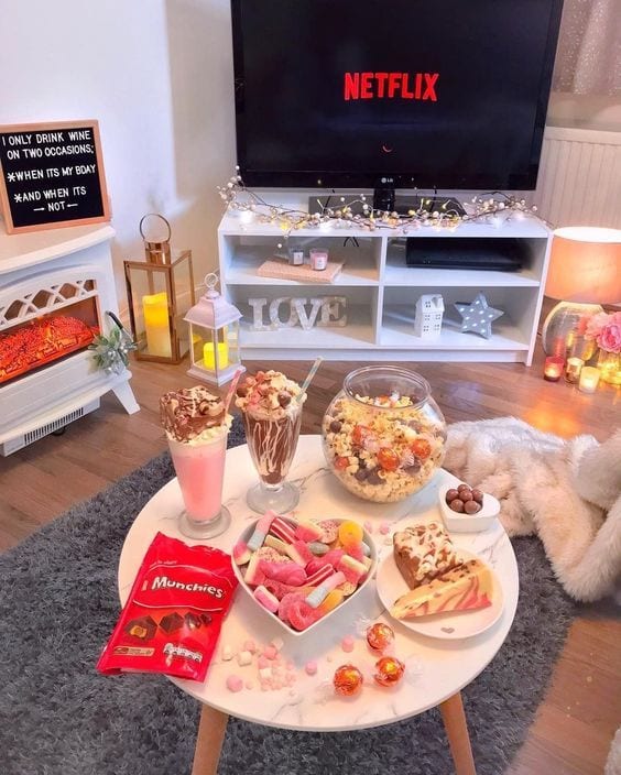 Mesa de centro com potes de doces como fini, chocolate, milkshake, e pipoca e a tv ligada com o logo da Netflix.