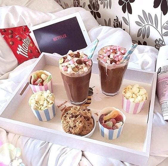 Bandeja branca para comer na cama, com milkshake, pipoca, cookies e notebook atrás com Netflix.