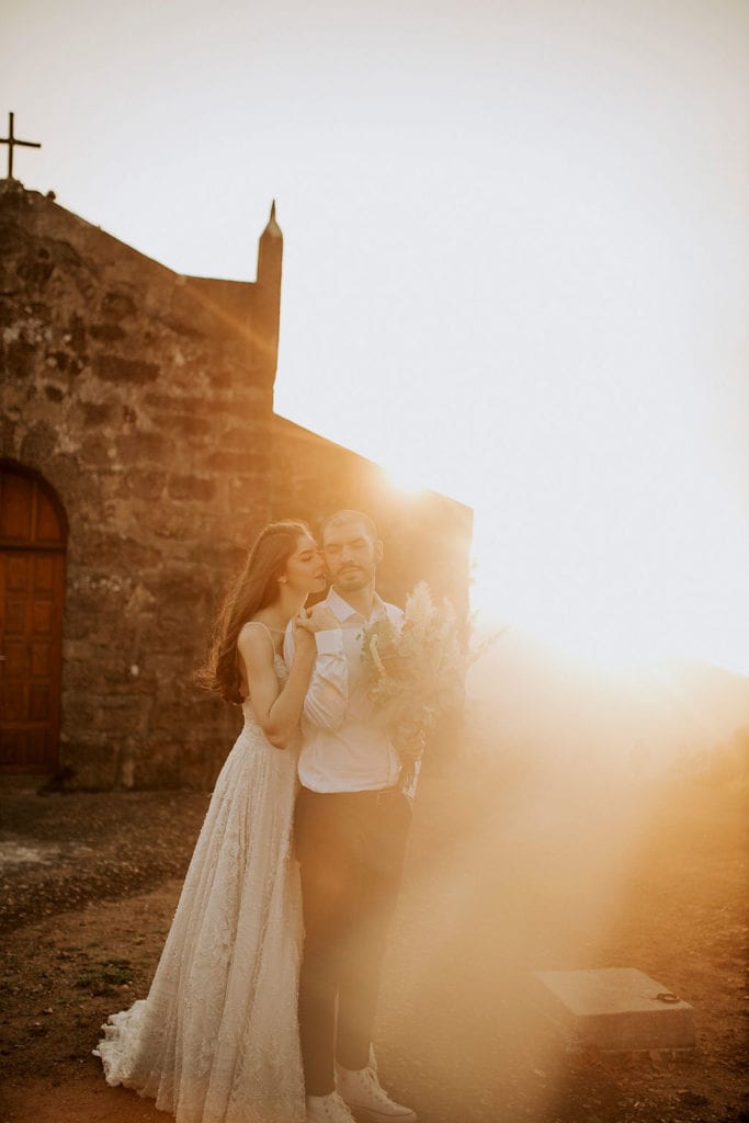 noivos posam no alto de um monte de frente para uma capela - fotos pós wedding