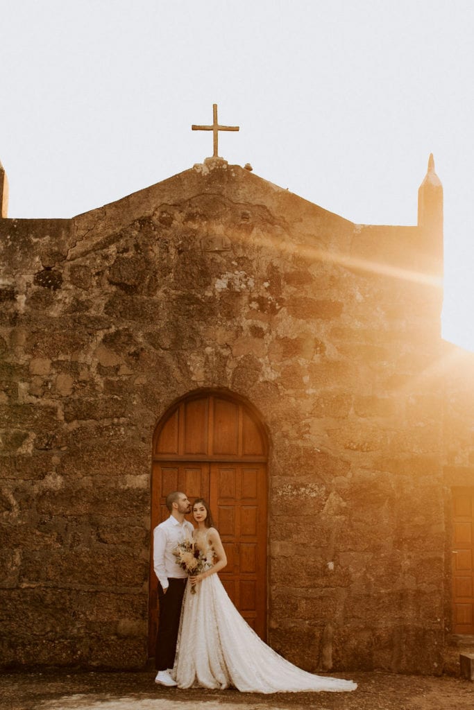 noivos posam no alto de um monte de frente para uma capela - fotos pós wedding
